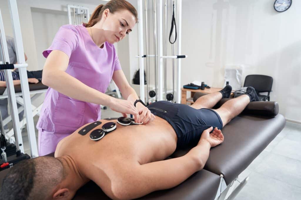 Was ist Elektrotherapie in der Physiotherapie? 1 achte Ein Mann, der sich in einer Physiotherapie-Praxis einer physiotherapeutischen Myostimulationsbehandlung (Elektrotherapie) unterzieht